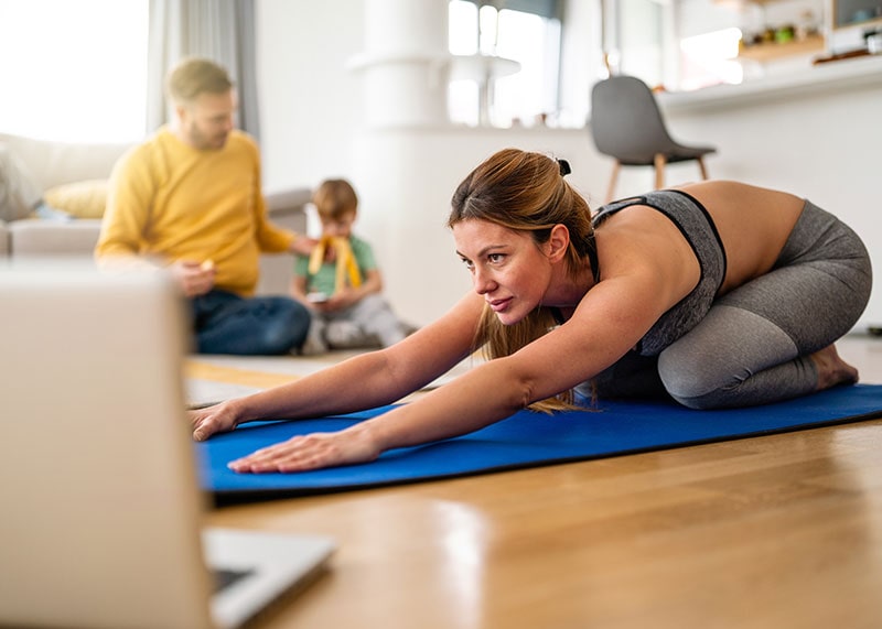 Mother exercising at home in living room father playing with kids in background